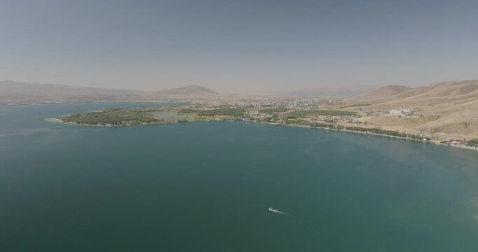 Aerial view of the calm waters of Lake Sevan, reflecting the hazy sky, contrasting with the arid landscape and the small boat leaving a white trail, Lake Sevan, Gegharkunik Province, Armenia.