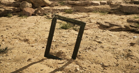 An empty black frame is positioned upright on sandy ground in a barren desert landscape. Surrounding rocks and sparse vegetation create a stark contrast against the frame.