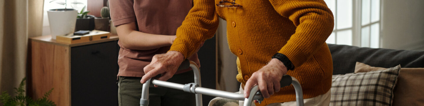 Senior Caucasian man using walker for support while young adult woman assisting him indoors, both standing close together, partial view focusing on upper bodies and hands