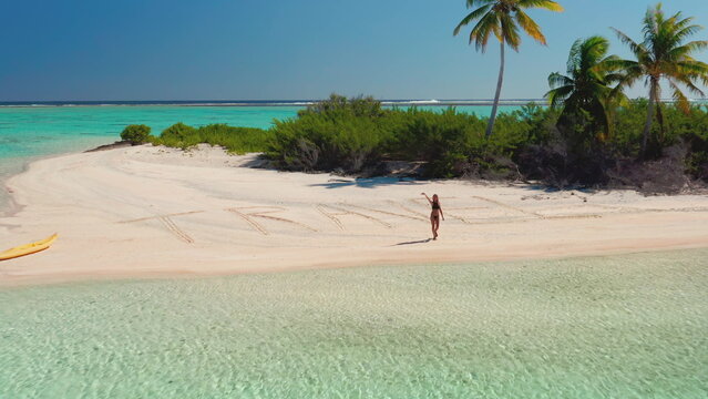 Aerial view of a tropical beach with a female tourist walking on the sand where the word travel is written. The turquoise water is crystal clear and a yellow kayak is waiting on the shore