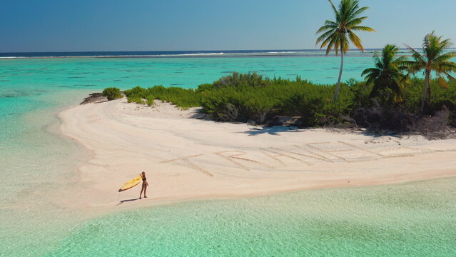 A person is walking on a sandy tropical beach, carrying a surfboard under their arm. The turquoise waters and palm trees can be seen in the background.