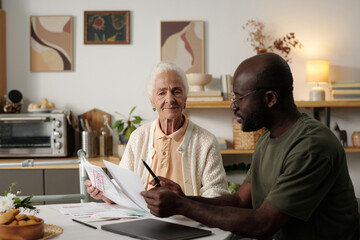 Senior Caucasian woman sitting at table reviewing documents with young Black man explaining paperwork, both focused on discussion in modern kitchen setting