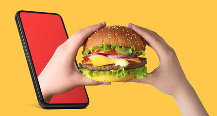 Food ordering and delivery service. Woman eating burger with one hand sticking out of mobile phone display against orange background, closeup