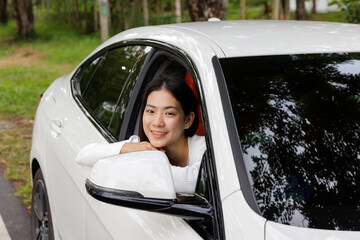 A young Asian cute girl happy smile from a car window sitting in a driver position beside a road while travel in the mountain and tropical forest trees