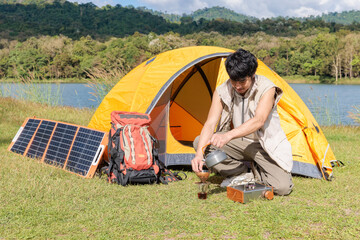 An Asian man sitting and prepare a cup of coffee near camping tent beside the lake and mountain in the sunshine morning with backpack and mobile solar cell
