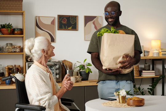 Senior Caucasian woman sitting in wheelchair smiling at young adult Black man holding paper grocery bag with vegetables, standing in kitchen engaging in friendly conversation - Powered by Adobe