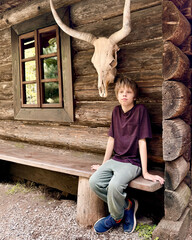 A teenager sits near a log house in a village, with an animal skull above his head.