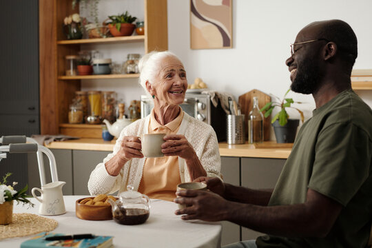 Senior Caucasian woman smiling while holding mug and sitting at table with young Black man, both engaging in friendly conversation in kitchen setting, enjoying hot drinks together