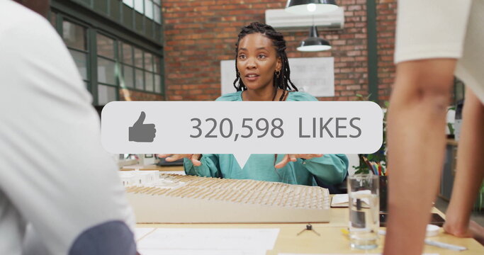 Presenting woman in teal blouse gesturing at architectural model on office desk, with likes overlay