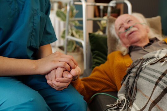 Senior Caucasian man lying in bed with oxygen tube holding hands with young adult woman caregiver, showing support and comfort in healthcare setting, walker visible in background