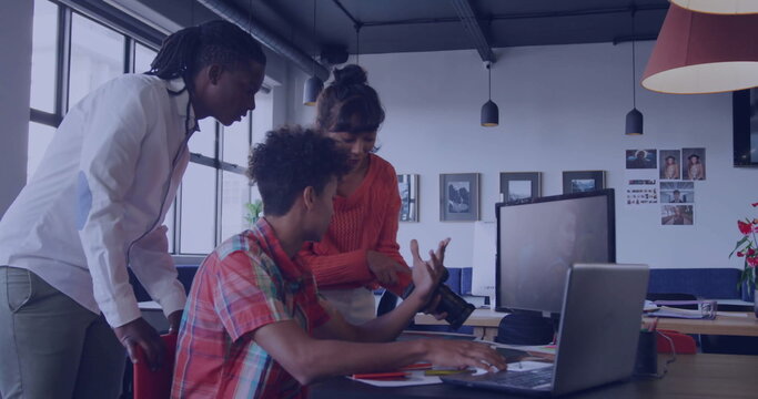 Pointing coworkers discussing DSLR camera lens and typing on laptop in office, with pendant lights