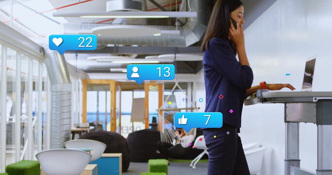 Blazer-clad woman using laptop and smartphone in office lounge, with social media icons, copy space - Powered by Adobe