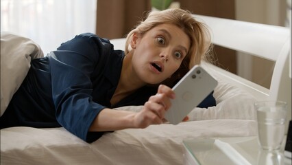 Shocked worried Caucasian woman overslept waking up bed looking mobile phone late in bedroom girl...