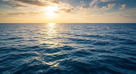 Fototapeta premium The sun shines brightly over a calm blue ocean with small waves under a partly cloudy sky at sunset.