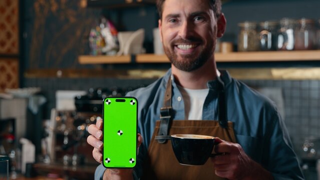 Glad Caucasian man barista small business entrepreneur male demonstrating mobile phone green screen smartphone application in cafe employee guy worker hold coffee drink cup restaurant online app tech