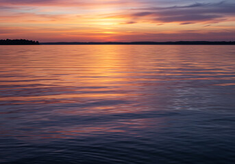 Obraz premium Sunset over calm water reflecting orange and purple hues, with a distant treeline silhouette on the horizon.