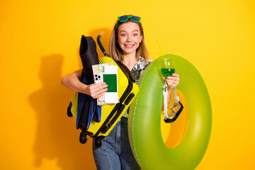 Cheerful young woman holding vacation essentials with vibrant yellow background expressing summer holiday excitement