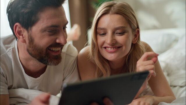 Family laughing Caucasian couple pair typing tablet shopping online in bedroom morning married woman discussing app wife man using technology Internet husband order food scroll social media together