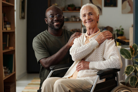 Portrait of senior Caucasian woman sitting in wheelchair with disability smiling at camera, while young Black man standing behind her gently placing hands on her shoulders
