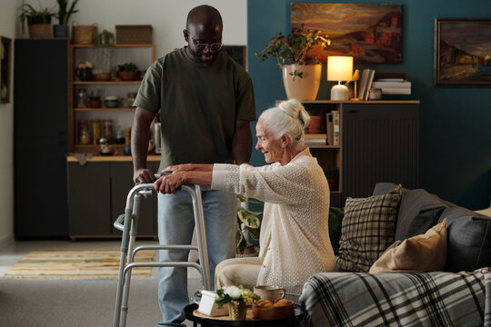 Senior Caucasian woman using walker assisted by Black man standing nearby in living room, both focusing on mobility support, elderly woman holding walker while man offering help