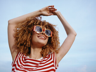 Joyful woman with curly hair wearing oversized sunglasses and a red striped shirt raises her arms against a bright blue sky, radiating carefree summer energy.