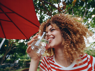 Bright outdoor scene features a joyful woman with curly hair sipping water from a glass under a red canopy, surrounded by lush greenery and warm sunlight.