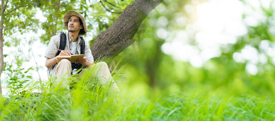 Asian man birdwatching with binoculars and notebook, studying wildlife in nature.