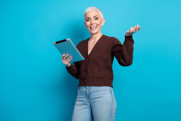 Confident senior woman with tablet, making presenting gesture, standing against vibrant blue...