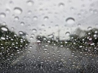 A close-up shot of raindrops on a car windshield on a rainy day. The water droplets vary in size and shape, with slight reflections. The background shows a blurred view of a road and trees, creating a