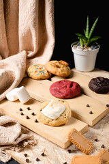 Stack of assorted gourmet cookies on wooden boards—red velvet, chocolate, sprinkle and marshmallow—styled in studio with black background; perfect for bakery promos, menu covers, and ads.