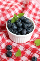 There is a small white bowl of blueberries on a checkered napkin.