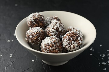 Tasty chocolate candies with nuts and coconut flakes in bowl on black table, closeup