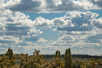 Rural landscape with clouds where large white with bluish cumulus clouds hang in blue sky above rural landscape with field and trees in the foreground in early autumn.