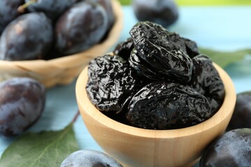 Dried prunes in bowl, leaves and fresh plums on light blue wooden table, closeup