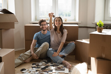 Overjoyed couple homeowners sitting on floor among boxes showing keys