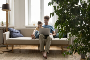 Caring young dad using notebook on sofa with little daughter