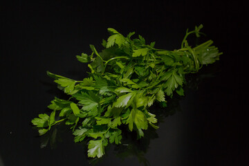 The photo shows a bunch of fresh green parsley lying on a dark, reflective surface. The bunch is neatly tied with a thin sprig. The black background highlights the rich color of the greenery.