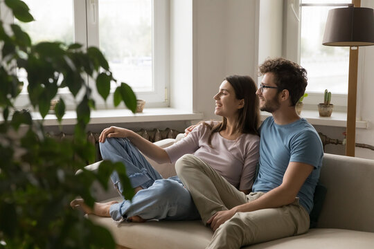 Young couple hug on sofa look aside with peaceful smiles