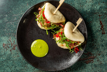 Two Asian-style bao buns with grilled meat, vegetables, and chili slices on a dark plate, served with green sauce. Modern food styling, top view, vibrant colors © pavel siamionov