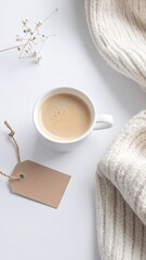 A white coffee cup filled with light brown coffee sits atop a white surface.  A beige knit sweater and dried flowers add a cozy touch