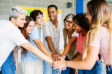 Diverse group of young high school students stacking hands with middle age female teacher in classroom showing unity, support and cooperation. Education concept
