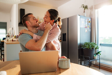 Young couple enjoys a tender moment together at their kitchen table, embracing affectionately with a laptop and coffee nearby, creating a warm and intimate scene of domestic bliss