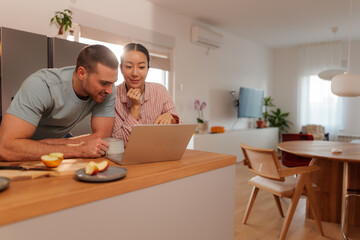 Fototapeta premium Young multi ethnic couple enjoying morning coffee while browsing the internet on a laptop in their modern kitchen, happily planning their day together in a cozy, bright space