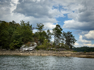 Laurel River Lake's forest trees growing along a rocky shoreline beside calm rippling lake water. Overhead dramatic clouds with patches of blue sky create a moody and peaceful atmosphere.