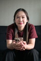 Fototapeta premium Young Thai woman sitting on sofa using smartphone at home, lifestyle portrait