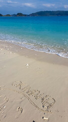 Word Okinawa is written in the wet sand on a beautiful beach, with gentle waves from the turquoise ocean washing ashore. Personal memento from a tropical vacation in Japan, surrounded by footprints.