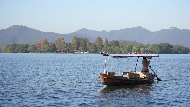 A wooden boat with a person rowing on West Lake, surrounded by distant mountains and calm water reflections