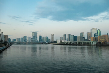 Naklejka premium The modern skyline of Tokyo rises above the calm Sumida River at dusk, where a bridge spans the water under a soft, cloudy sky, capturing the tranquil beauty of Japan's bustling capital city.