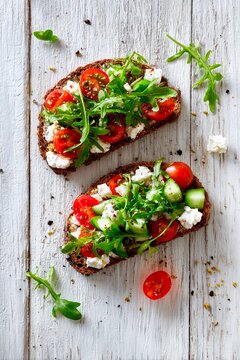 Two slices of whole grain bread are generously layered with fresh arugula, cherry tomatoes, cucumber, and soft cheese, creating a healthy and vibrant meal on a rustic wooden surface