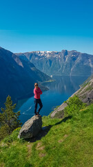 Naklejka premium A triumphant female hiker stands confidently on a rock at the Kjeåsen viewpoint, overlooking a Norwegian fjord. Her pose against the vast mountain landscape celebrates adventure and outdoor travel.